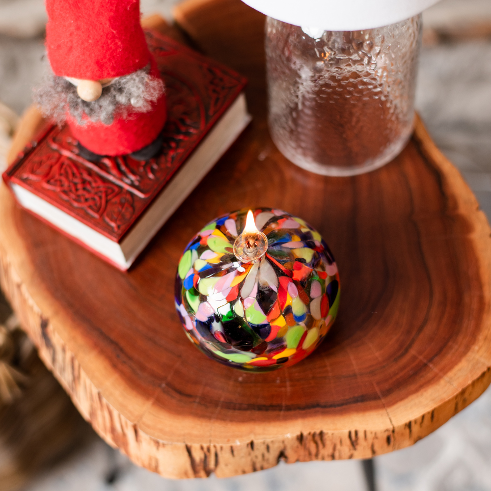 Lit round glass ball with flat base and wick opaque pink, blue, yellow, purple, opaque green, opaque yellow, lime and opaque white glass mosaiced around the entire surface on a wooden table with a red gnome to the left and behind