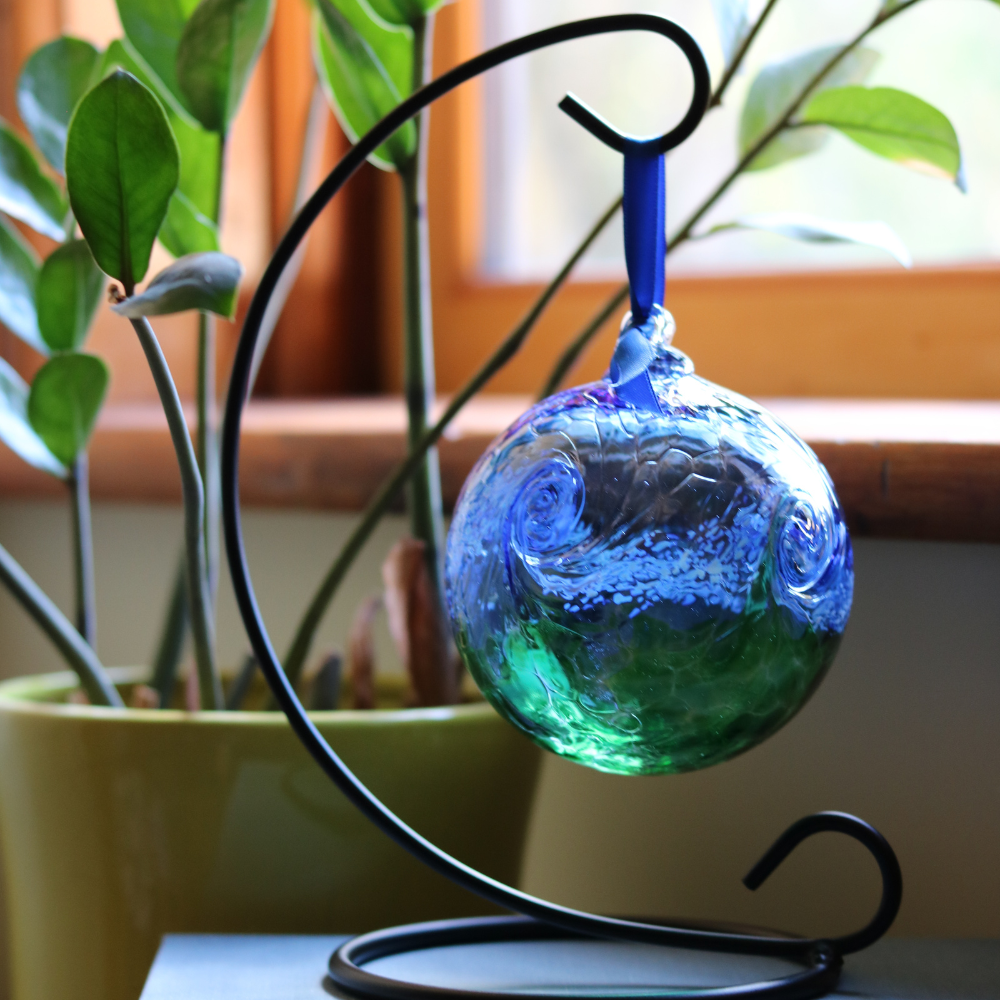 Glass orb with blue on top, white swirled across the middle, and green on the bottom hanging on a black curved ornament holder in front of a green plant and window.