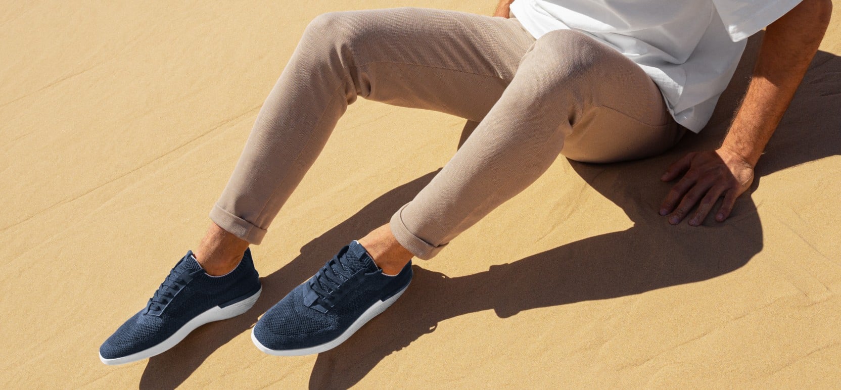 Waist down view of man sitting wearing supremeknit trainer navy with a sand background