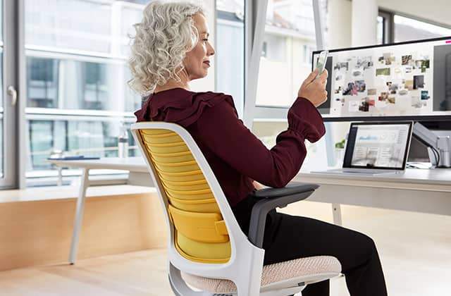 Mujer sentada en silla de oficina Steelcase Serie 1 en Níquel/Canario, foco en el respaldo, en una oficina.