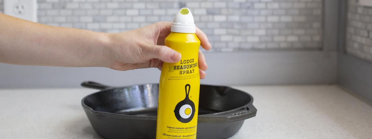 A person sets the bottle of Seasoning Spray onto the countertop in front of a Lodge Cast Iron Skillet.