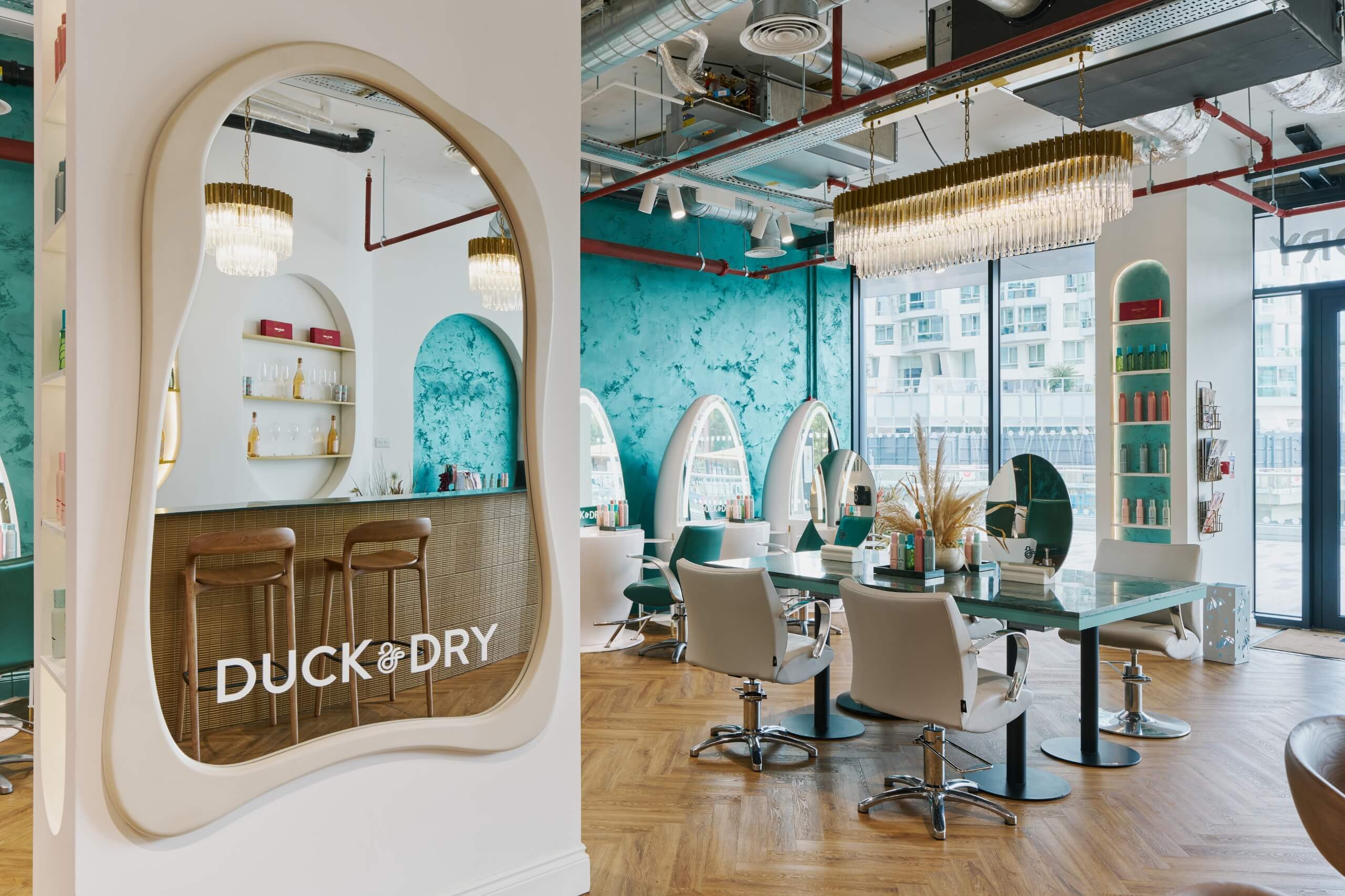 Duck & Dry Battersea blow dry salon interior with teal textured walls, arched illuminated mirror stations, gold chandeliers and styling chairs, with the Duck & Dry logo on a bar area to the left.