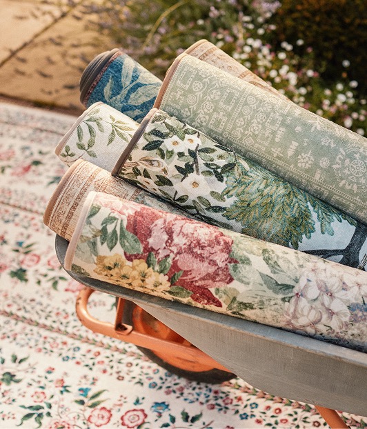 Lot of runners from the Highgrove by Sanderson x Hug Rug collection rolled up and placed in a wheelbarrow, with a rug on the ground underneath. 