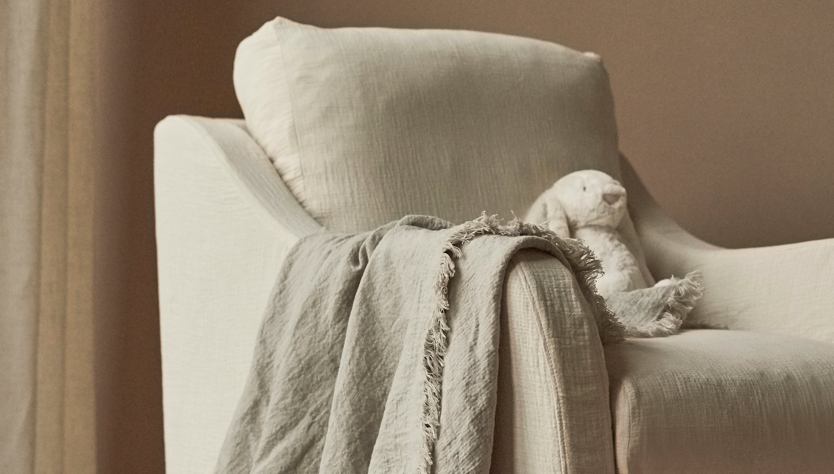 Close-up view of Esme Glider Chair in Corn Silk, a light beige Washed Cotton Linen with a blanket and toy on the seat