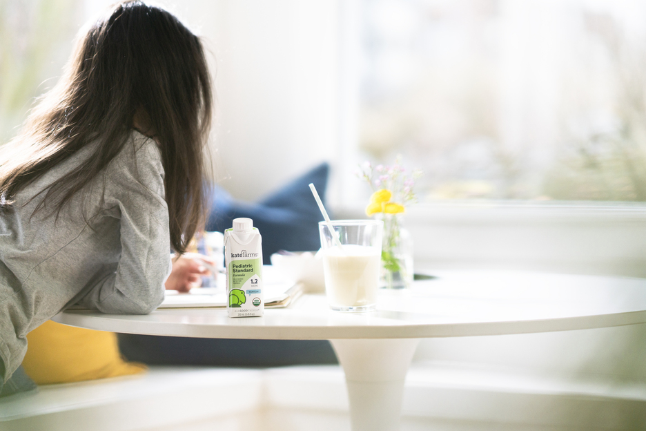 A child is doing homework at a table with a carton of Kate Farms Pediatric Standard 1.2 formula sitting next to the shake poured into a tall glass with a straw.