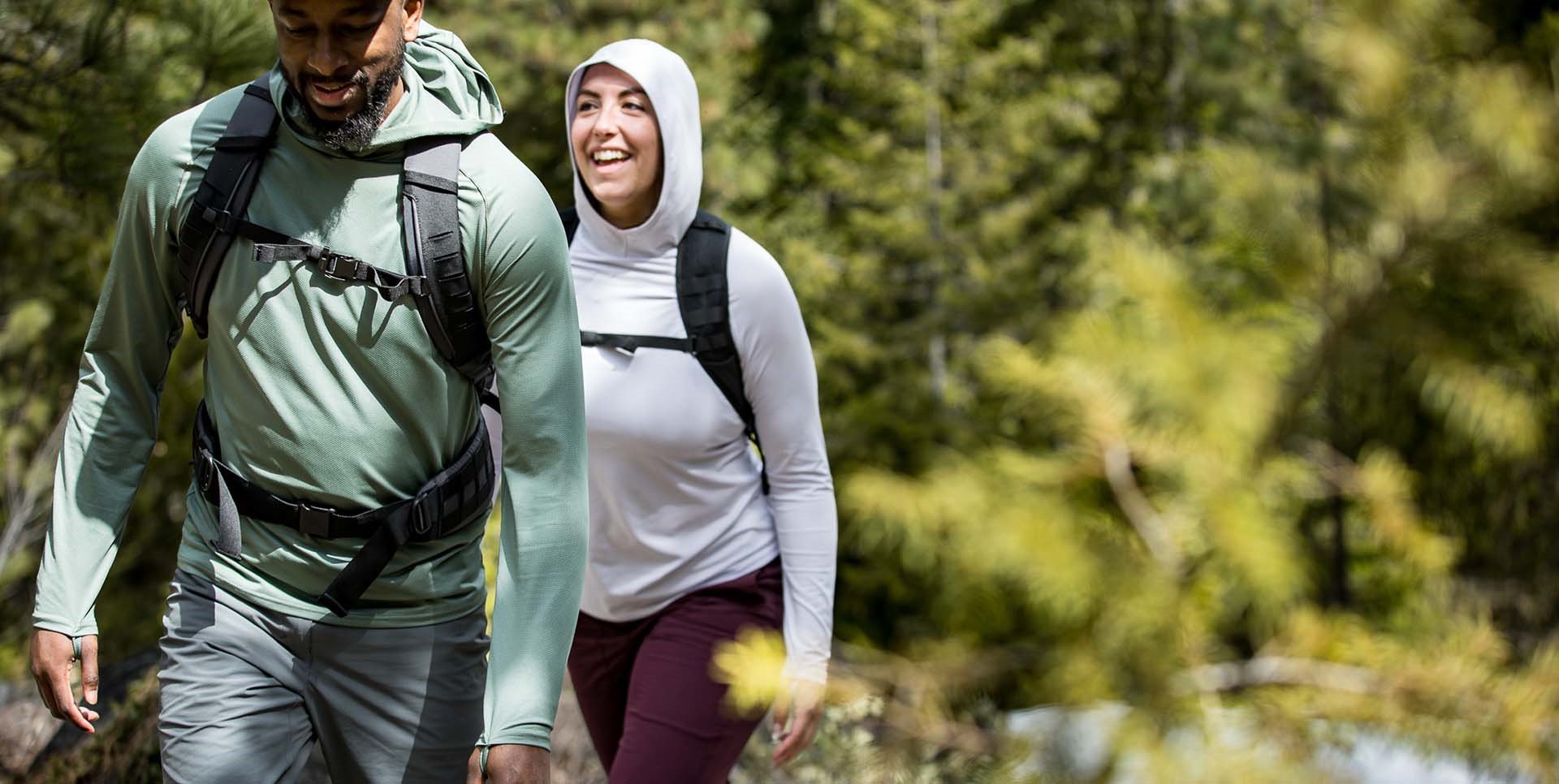 Man wearing Dark Mint Geo T Hooded Ls Shirt and Woman wearing Lilac Grey Geo T Hooded LS Shirt hiking through the sunny forest