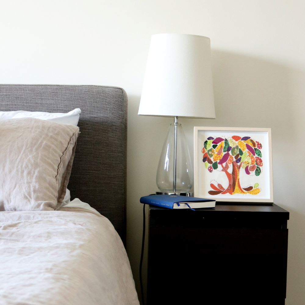 orange, brown, cranberry, yellow, green and blue fabric tree on a white background in a wood frame. Displayed on a black side table beside a blue notebook and bed with grey headboard and white linens. White lamp in the background