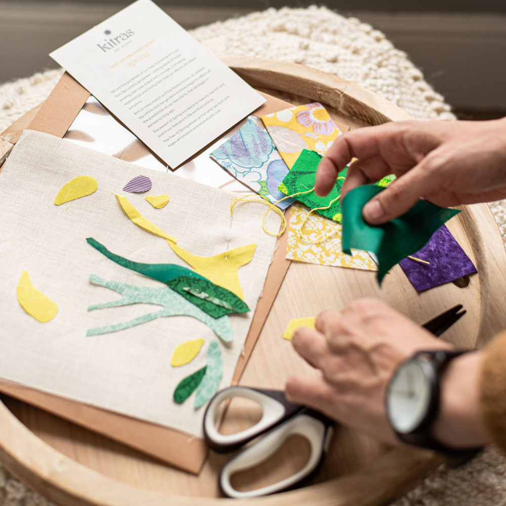 Partially completed Spring slow stitch kit. green and yellow cut fabrics on a white canvas. Hands holding a green piece of fabric, reaching for scissors. Tree of Spring story on a white card at the top