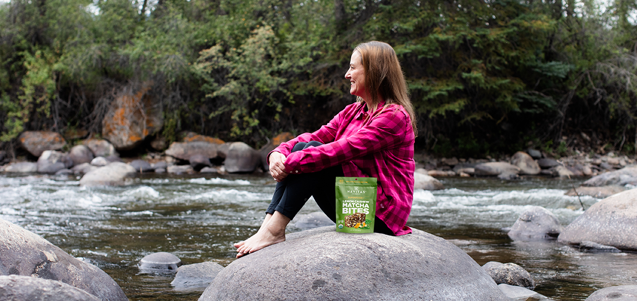Person sitting on a rock by a river, next to a package of Navitas Organics Superfood+ Lemon Cashew Matcha Bites, surrounded by a forested area.