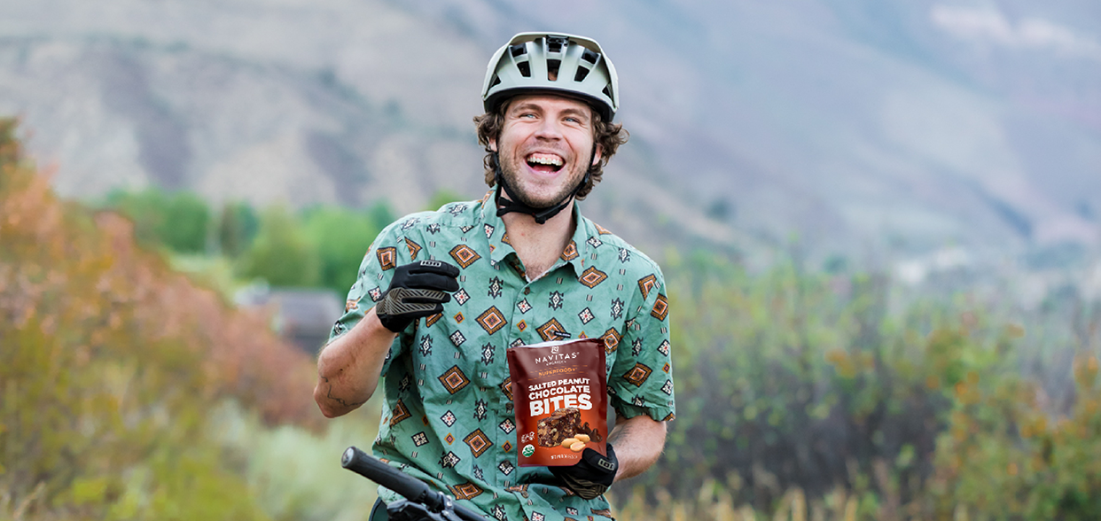Person on a bicycle holding a bag of Navitas Organics Salted Peanut Chocolate Bites, smiling, wearing a bike helmet and a patterned shirt, with a blurred natural landscape in the background.