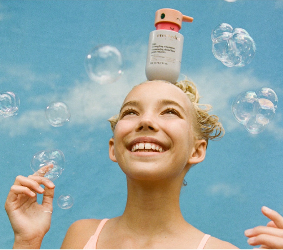Smiling child balancing a kids shampoo bottle on their head with bubbles around them.