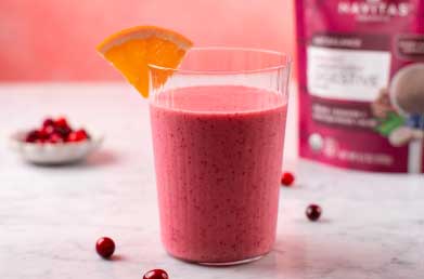 A glass of smoothie with a slice of orange on the rim, surrounded by scattered cranberries and a bag of Navitas Organics Superfood+ Digestive Blend in the background.