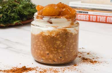 A jar of overnight oats topped with a dollop of apple puree and a sprinkle of cinnamon, placed on a white marble surface.