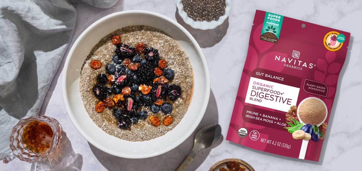 A bowl of chia seed pudding topped with blackberries and blueberries next to a honey dipper, with a package of Navitas Organics Superfood+ Digestive Blend visible in the background.