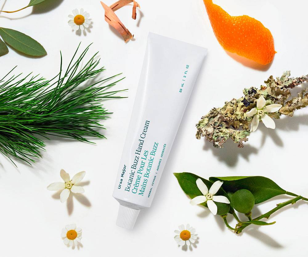 White aluminum tube of Ursa Major Botanic Buzz Hand Cream tube on a white background with nature ingredients around it.