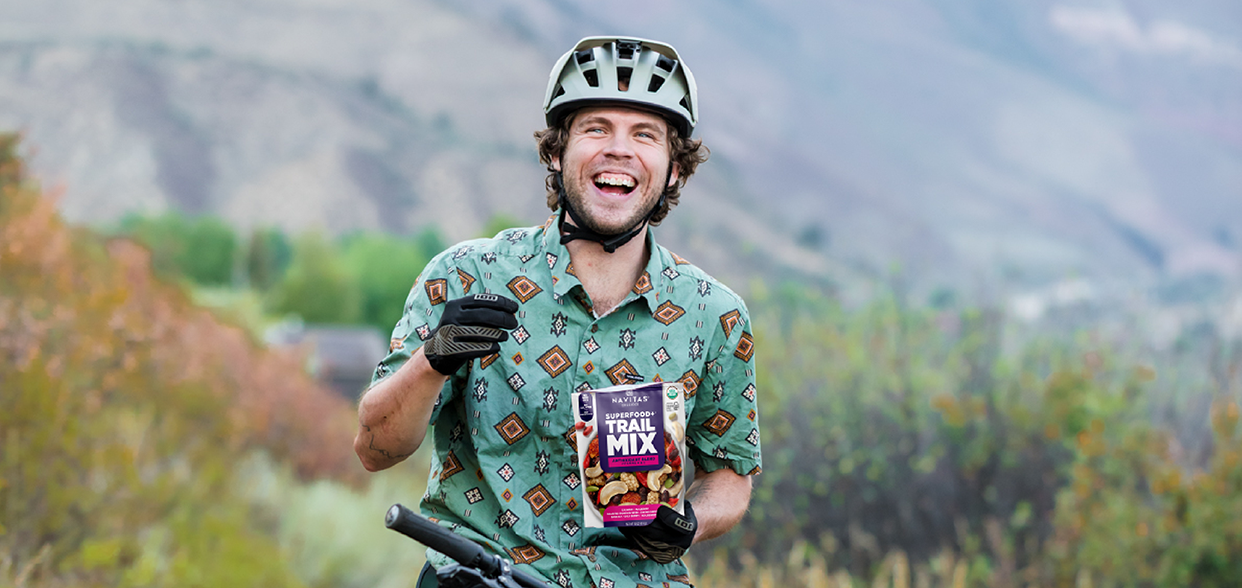 Person in a helmet and patterned shirt smiling while holding a bag of Navitas Organics Superfood+ Trail Mix Antioxidant Blend, standing with a bicycle in a natural, mountainous setting.