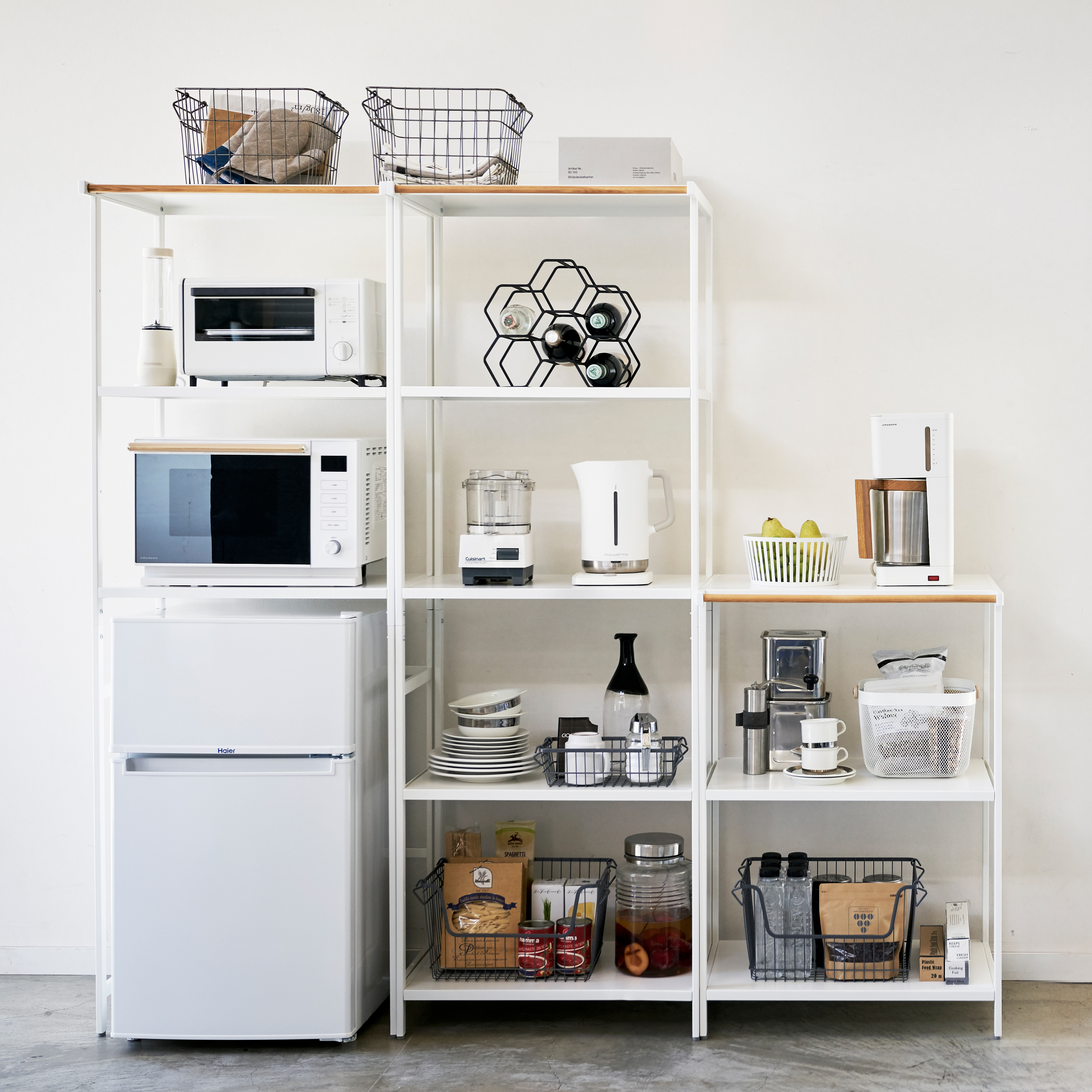 Three white minimalist shelving units with light wood tops stand against a white wall, filled with kitchen appliances, cookware, and pantry items in an organized display.