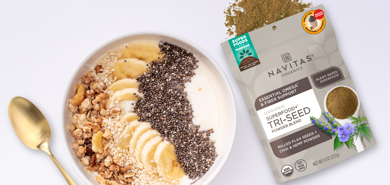 A smoothie bowl topped with granola, seeds, and banana slices surrounded by a gold spoon and a bag of Navitas Organics Superfood+ Tri-Seed Powder Blend.
