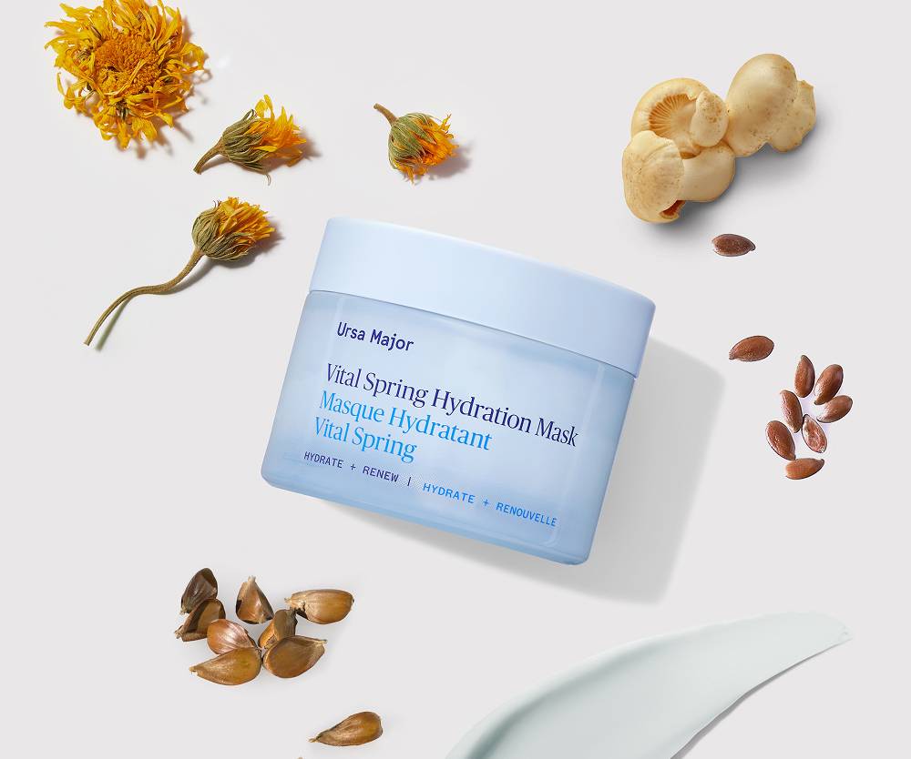 Blue jar of Ursa Major Vital Spring Hydration Mask, with botanical ingredients like flowers and mushrooms spread around it, on a white surface