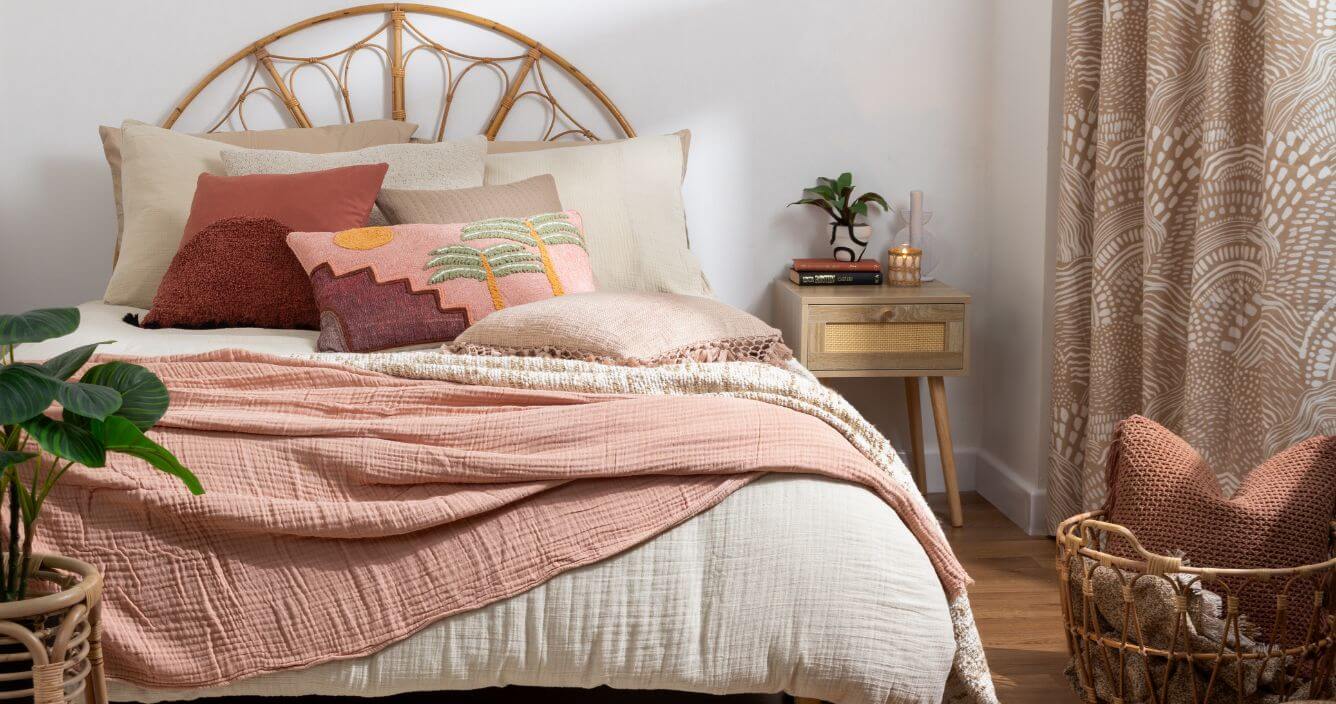 Neutral bed accessories layered in a boho style, including a beige duvet, red and pink cushions, and a clay pink throw.