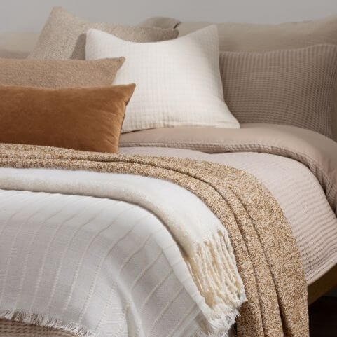 Neutral home decor layered on a minimalist bed, including a waffle linen duvet cover, white, beige and brown cushions, and textured throws.