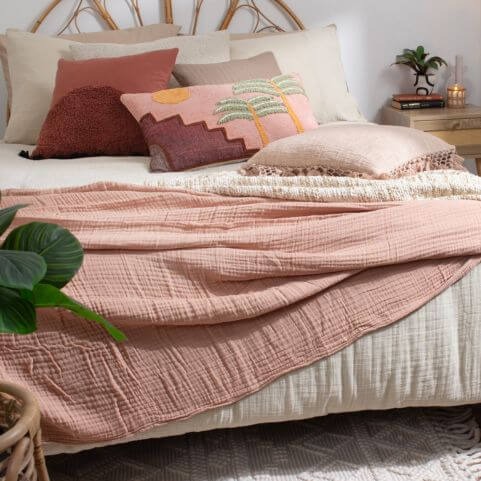 Neutral home decor in a boho style bedroom, including a natural muslin duvet set, pink and red scatter cushions, and a clay pink throw.