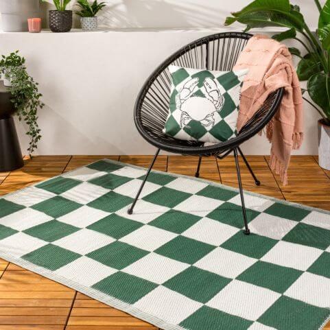 A polypropylene outdoor rug with a green and white checkerboard design, displayed on a wooden surface under garden furniture.