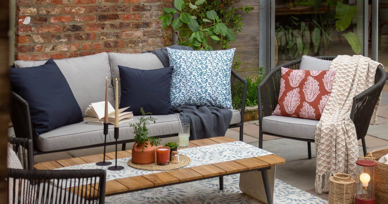 A small garden patio space decorated with a sofa, outdoor cushions, throws and an outdoor rug.