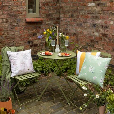 A small patio garden space decorated with a bistro set and floral outdoor cushions.