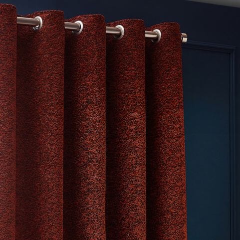 One panel of room darkening eyelet curtains in a copper red shade, hung on a metal curtain pole in front of a navy blue wall.