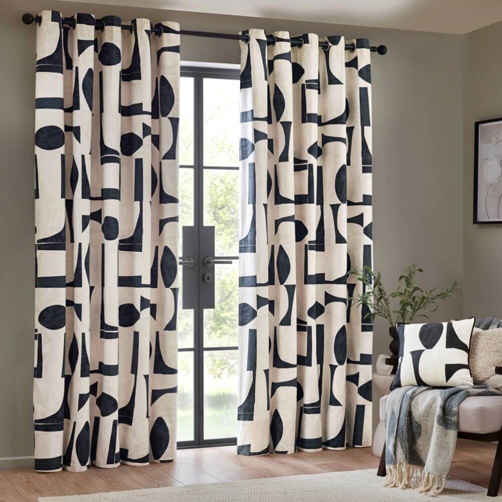 A set of 100% cotton, neutral curtains with an abstract geometric design in a dusky blue shade, hung on a black curtain pole in front of a grey wall.