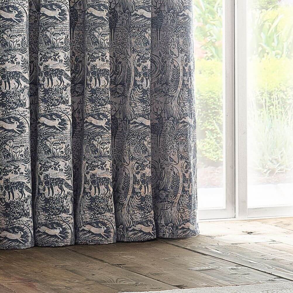 A close-up image of a curtain panel grazing a wooden floor. The curtains are neutral midnight blue in colour and feature a detailed design of woodland animals and foliage.