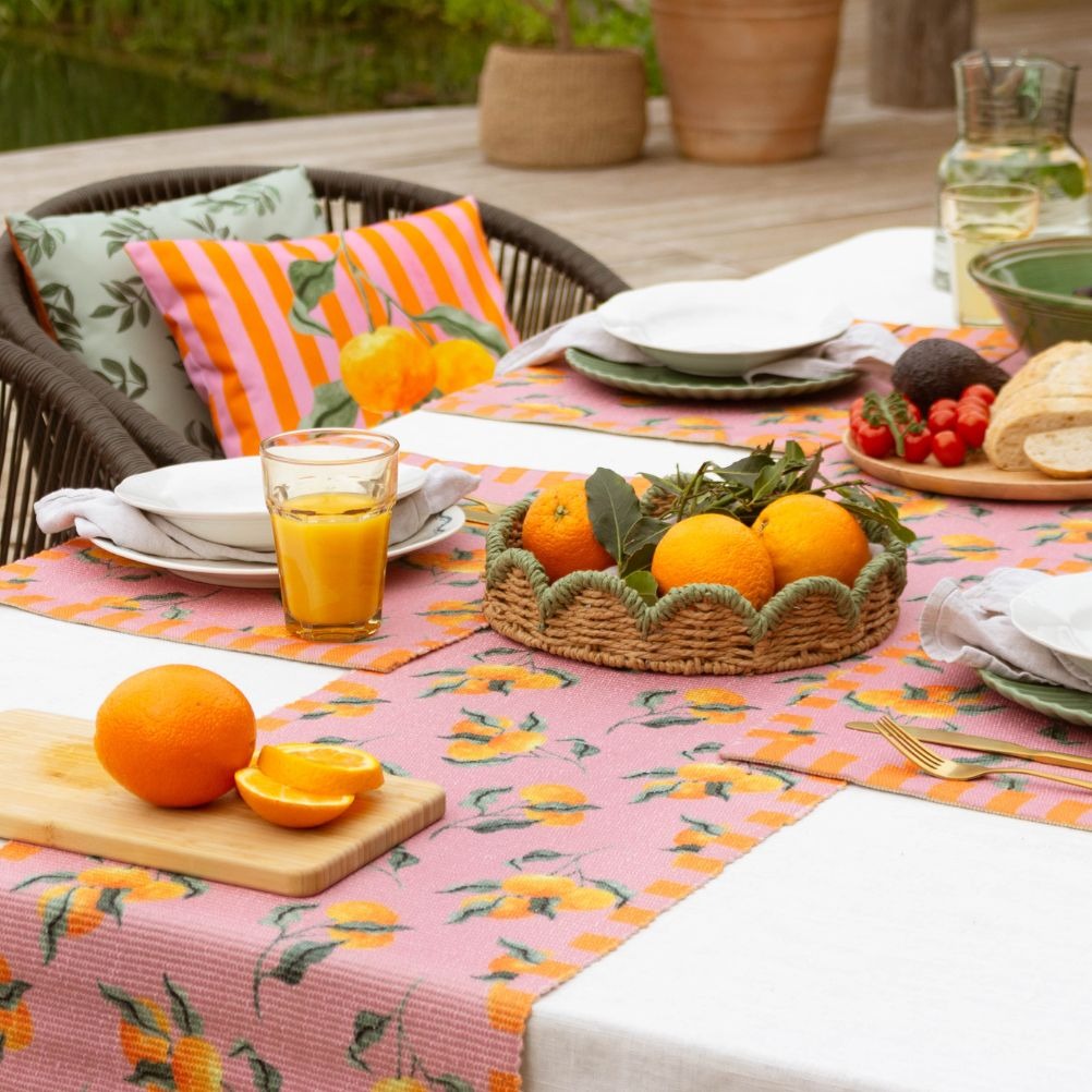 A colourful outdoor table set with orange-themed runners, placemats, plates, fruit, and drinks. 