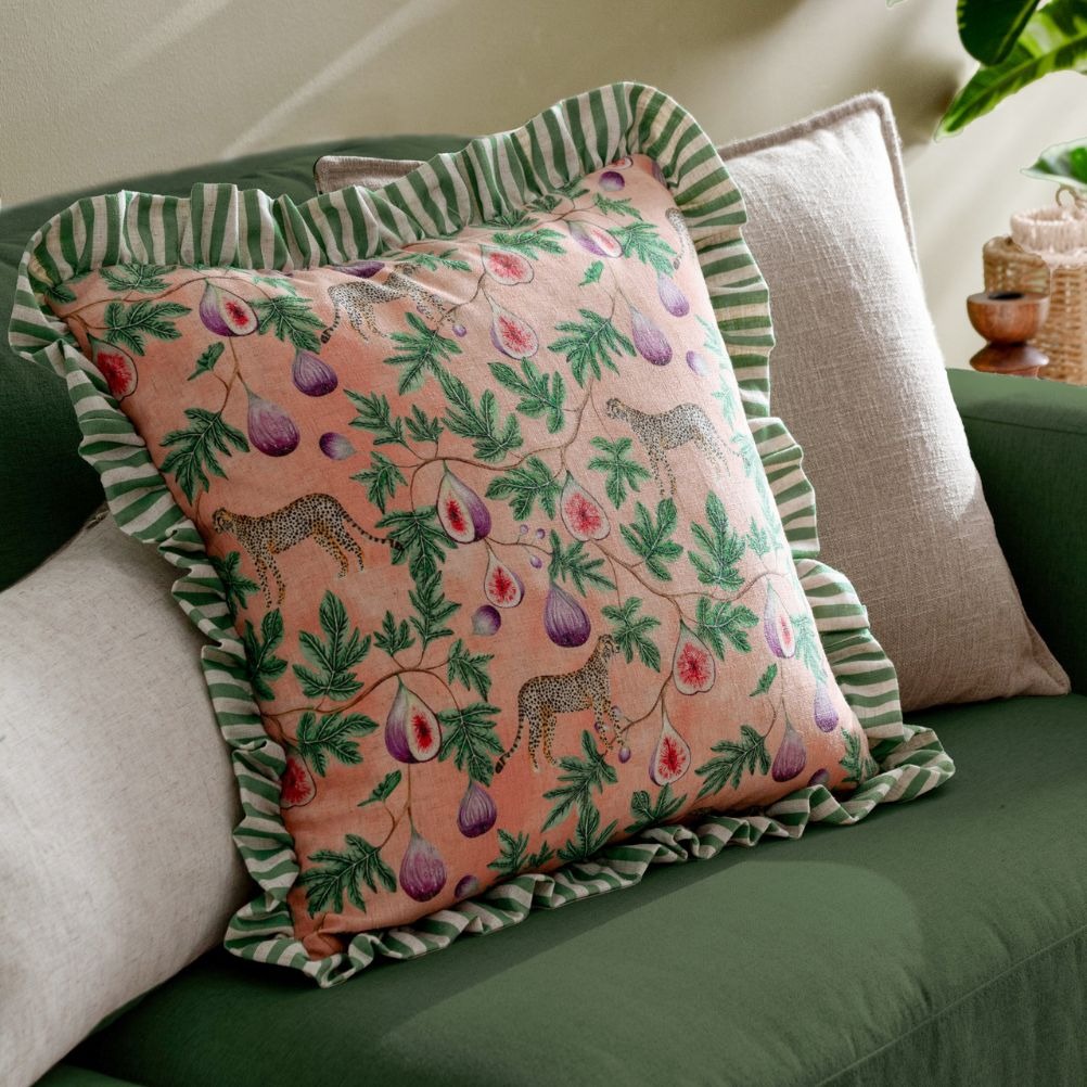 A decorative cushion with a pink pattern featuring leopards and figs, bordered with green striped ruffles. 