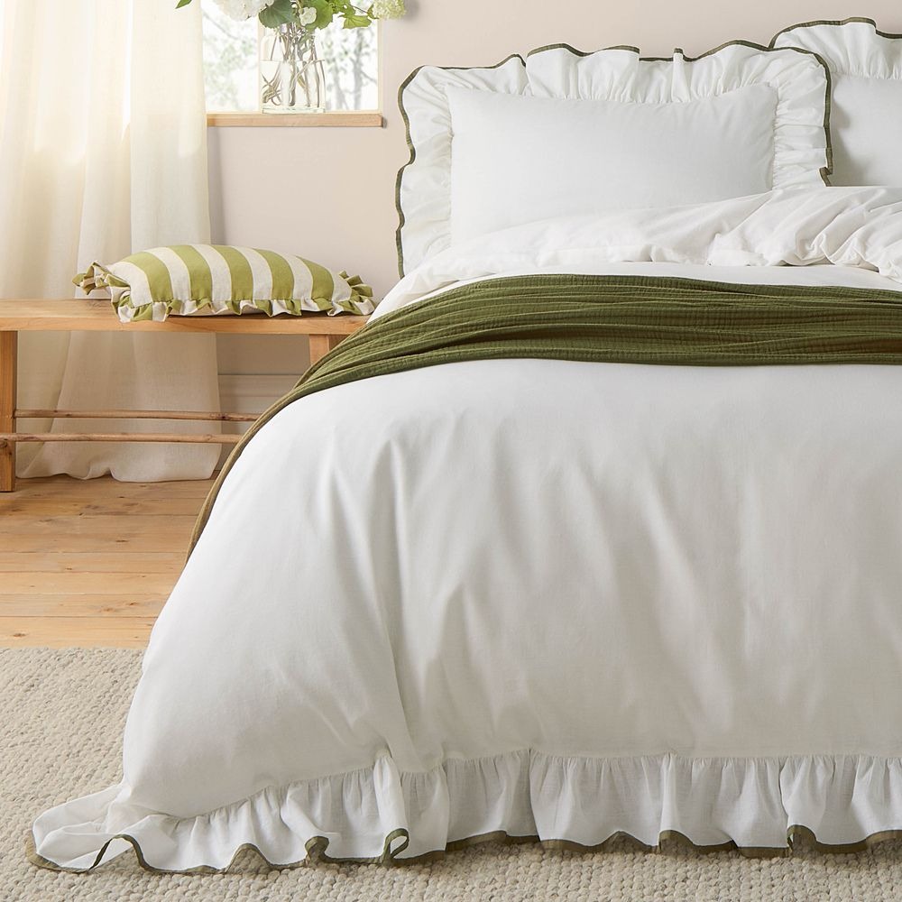 White frilly bedding with dark green trimmed edge, green throw and a cream and crean striped frilly cushion on a side table