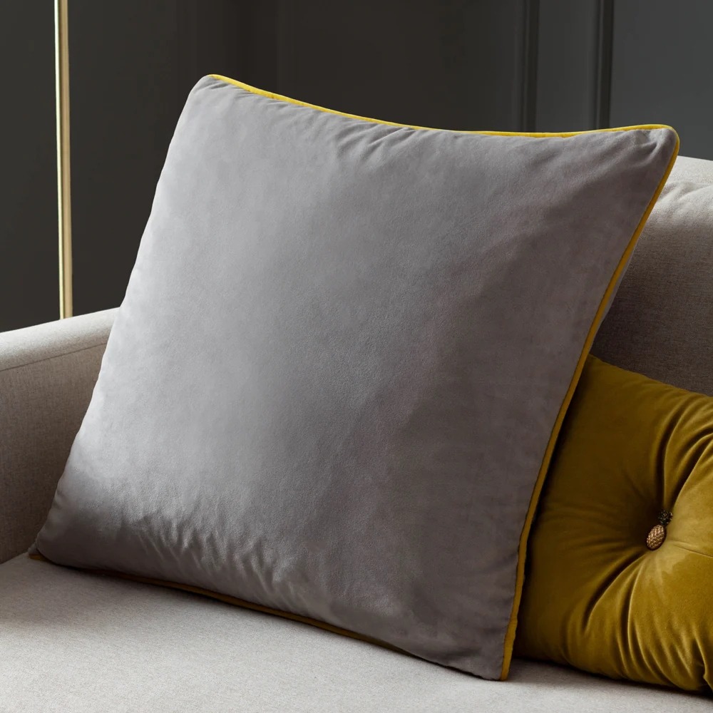 grey velvet cushion with a yellow piped edge in front of a small yellow cushion on a grey armchair.