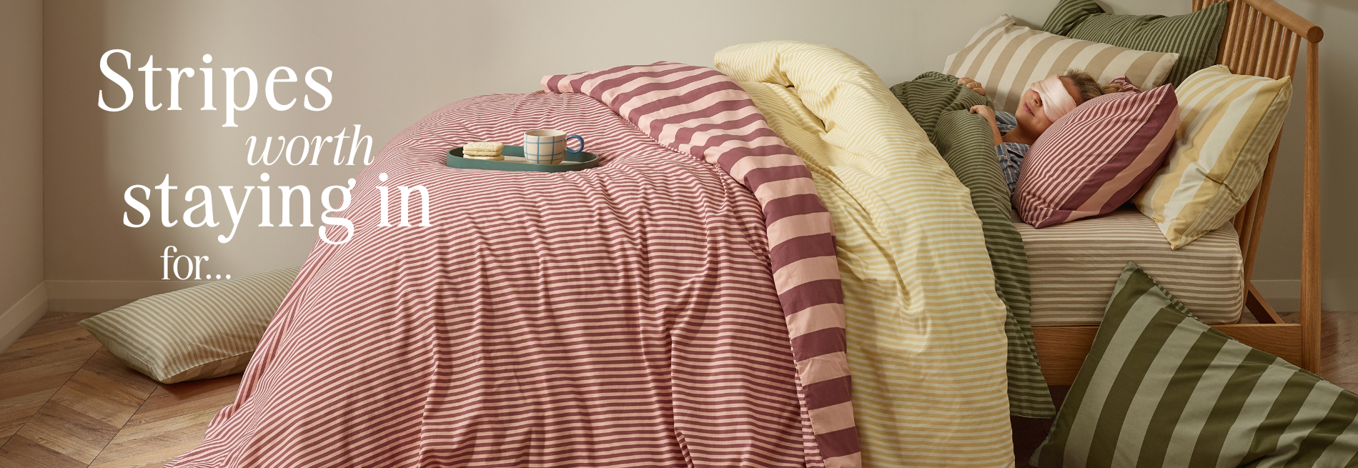 A wooden bed frame with three duvets on top, one with pink stripes, one with yellow stripes, and one with green stripes with matching pillowcases, and a girl sleeping in them with a pink silk eye mask on, with 