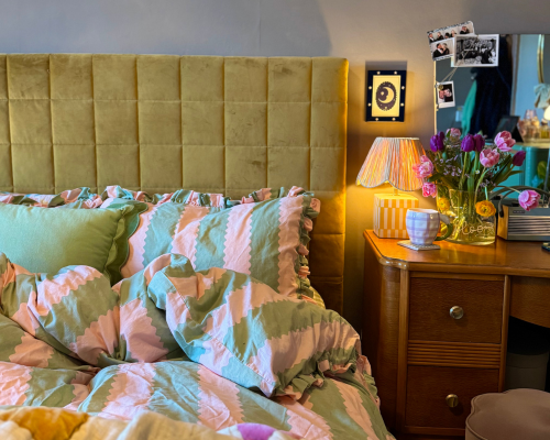 A pink and green wide stripe duvet cover with a green velvet cushion, next to a wooden bedside table with a warm lamp and flowers