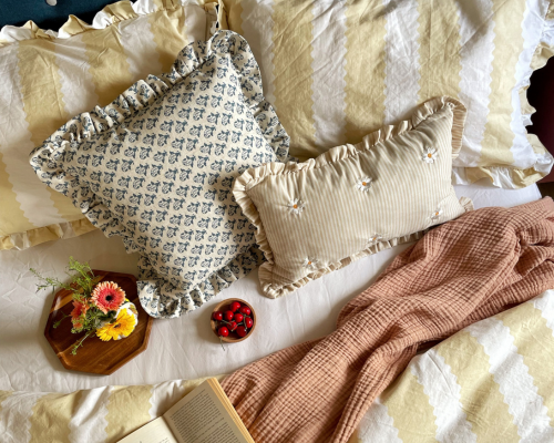 A butter yellow and white striped bedding set, with a yellow striped cushion and a blue floral cushion both with a frilled edge trim