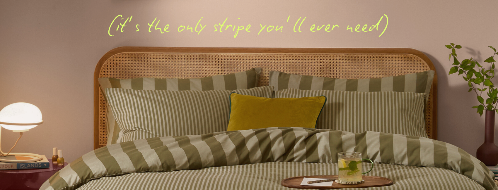 A green striped bedding, with a wider stripe on one side, and a narrow one on the other. There is a white lamp on a brown oak bedside table and a green plan on the other side.