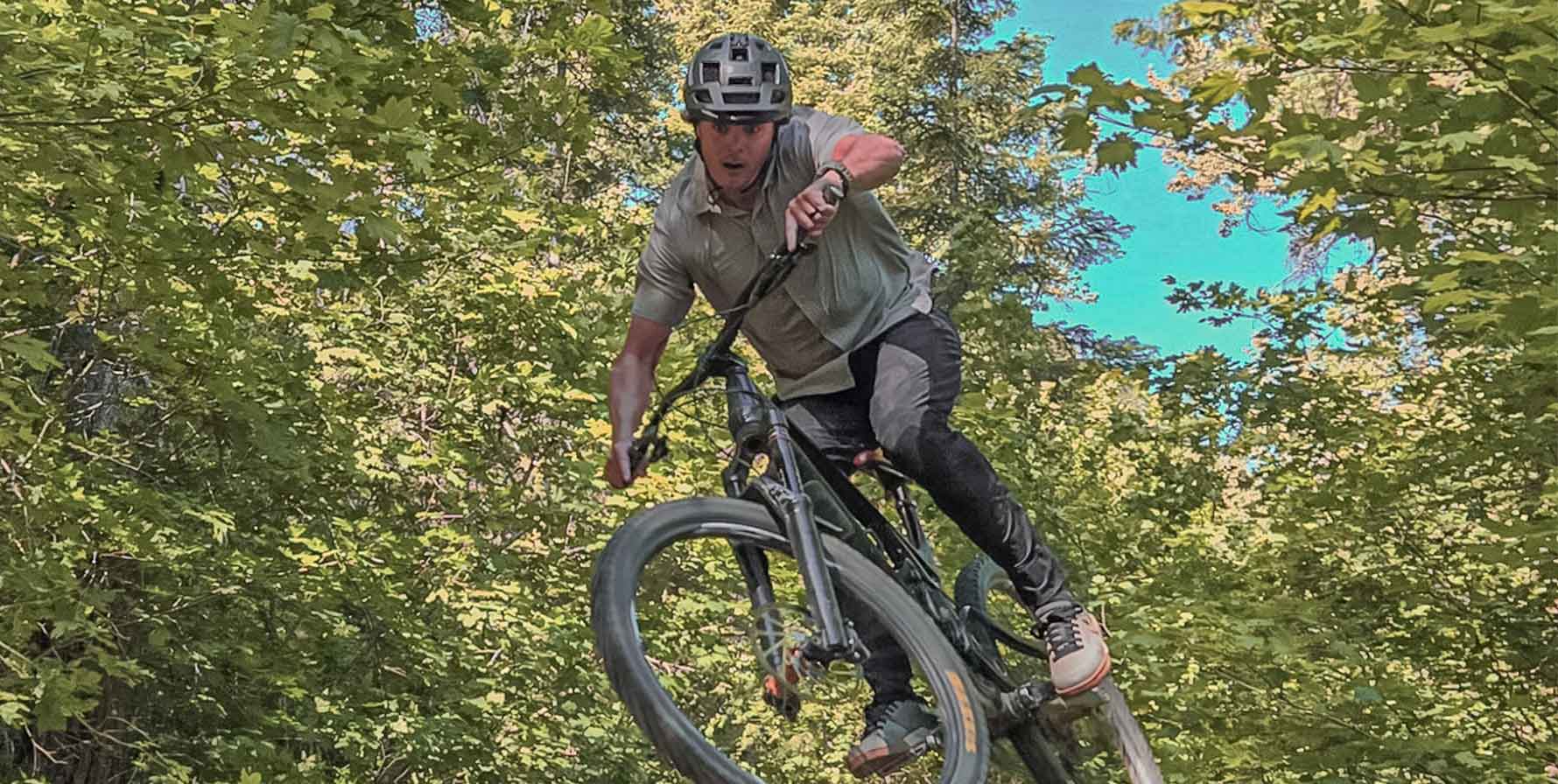 Man wearing the Moonstone FirstSun shirt and Nightbreak Camo Avid Pant while mountain biking in mid-air. Tree leaf filled background.