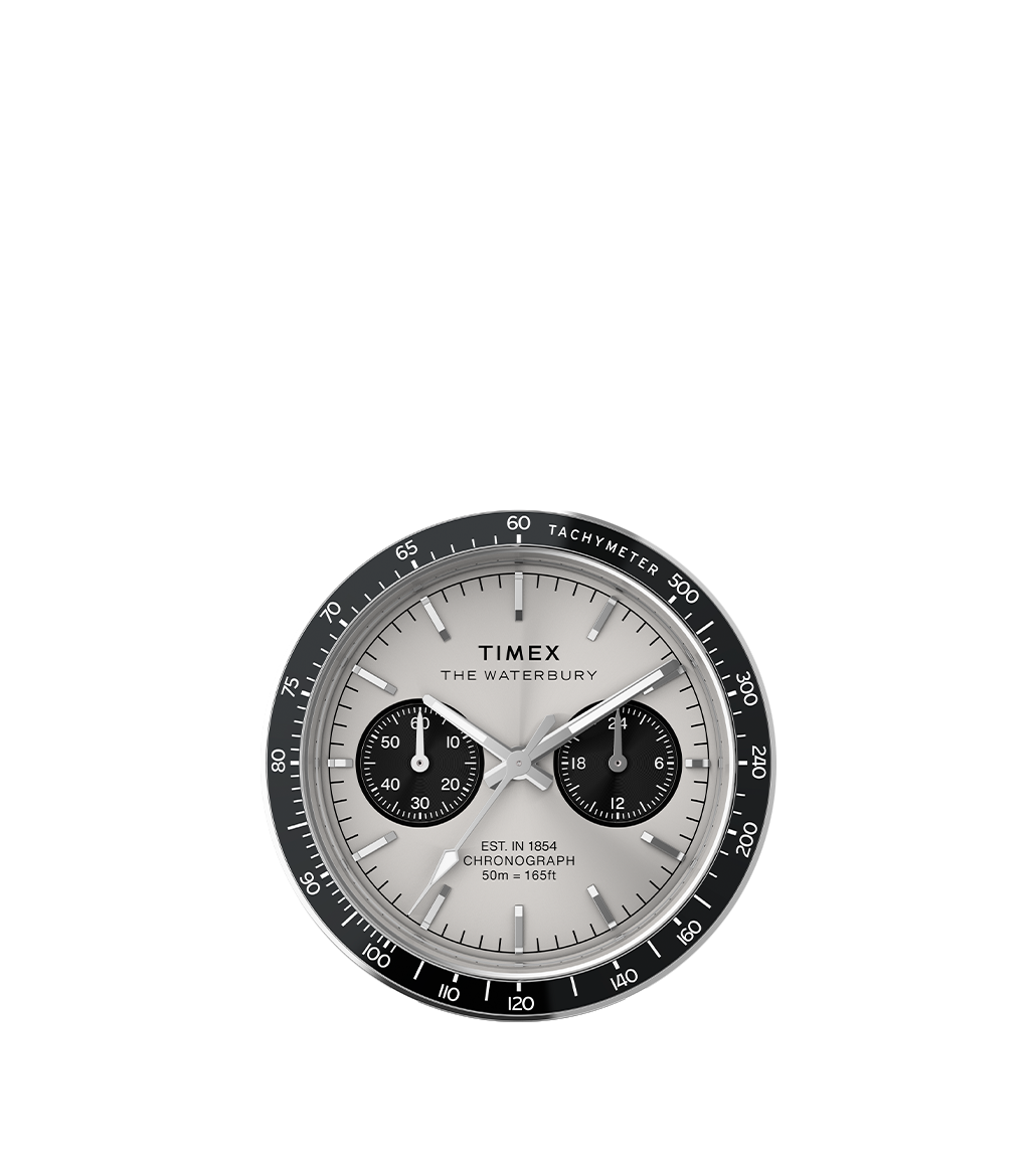 A close-up of the Waterbury Heritage Chronograph watch, showcasing its silver dial, black subdials, and black tachymeter bezel. The watch hands and markers are clearly visible, emphasizing its detailed craftsmanship.