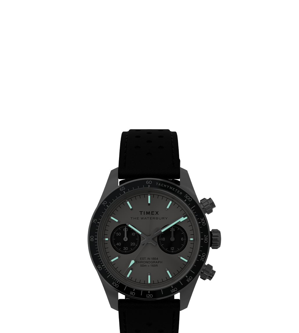 A luminous view of the Waterbury Heritage Chronograph in a dimly lit environment, highlighting its glow-in-the-dark hands and markers. The watch's sleek black strap and case contrast with the illuminated details.