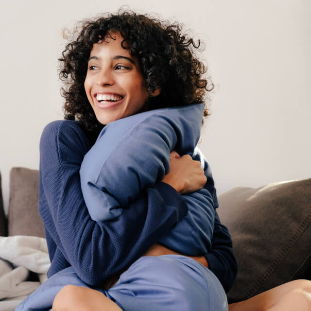 A woman sitting on her couch smiling and snuggling a pillow with the Slumber Cloud Essential Pillowcases