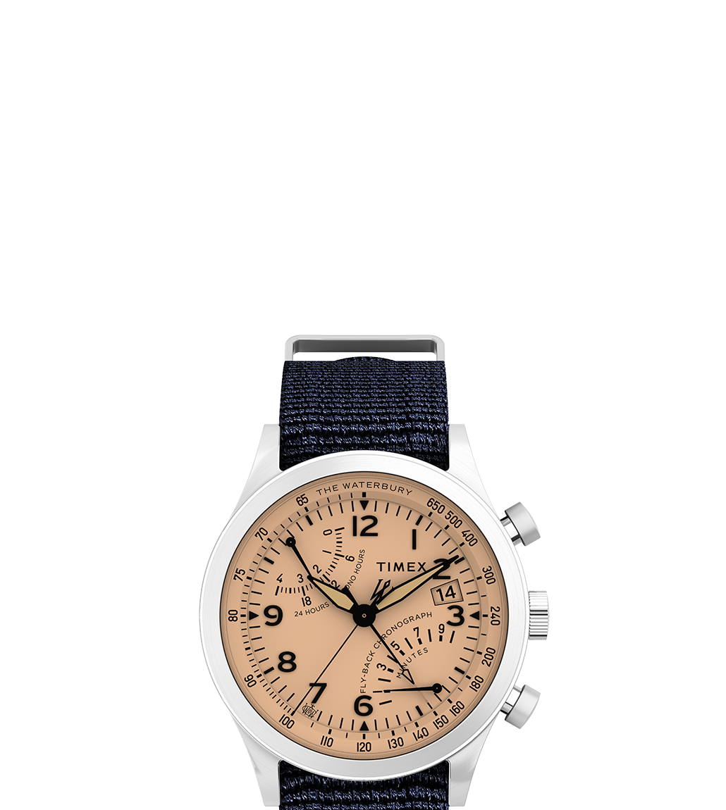 Front-facing product shot of the Waterbury Traditional Fly-back Chronograph against a white background. Highlights include a brushed stainless steel case, peachy-salmon dial with black Arabic numerals, chronograph sub-dials, a date window at 3 o’clock, and a textured navy-blue fabric strap.
