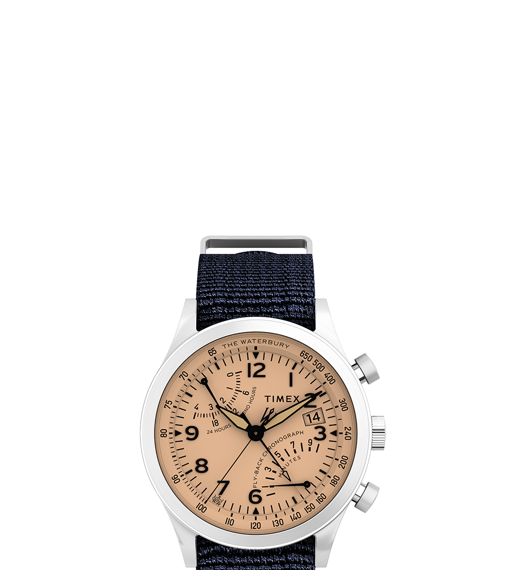 Front-facing product shot of the Waterbury Traditional Fly-back Chronograph against a white background. Highlights include a brushed stainless steel case, peachy-salmon dial with black Arabic numerals, chronograph sub-dials, a date window at 3 o’clock, and a textured navy-blue fabric strap.