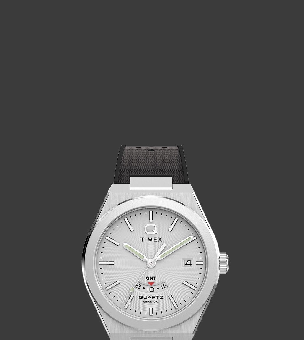 Front view of the Q Timex® Continental GMT watch with a silver-tone case and dial, black hour markers and hands, black strap with textured detail, and a GMT sub-dial located above 6 o’clock.