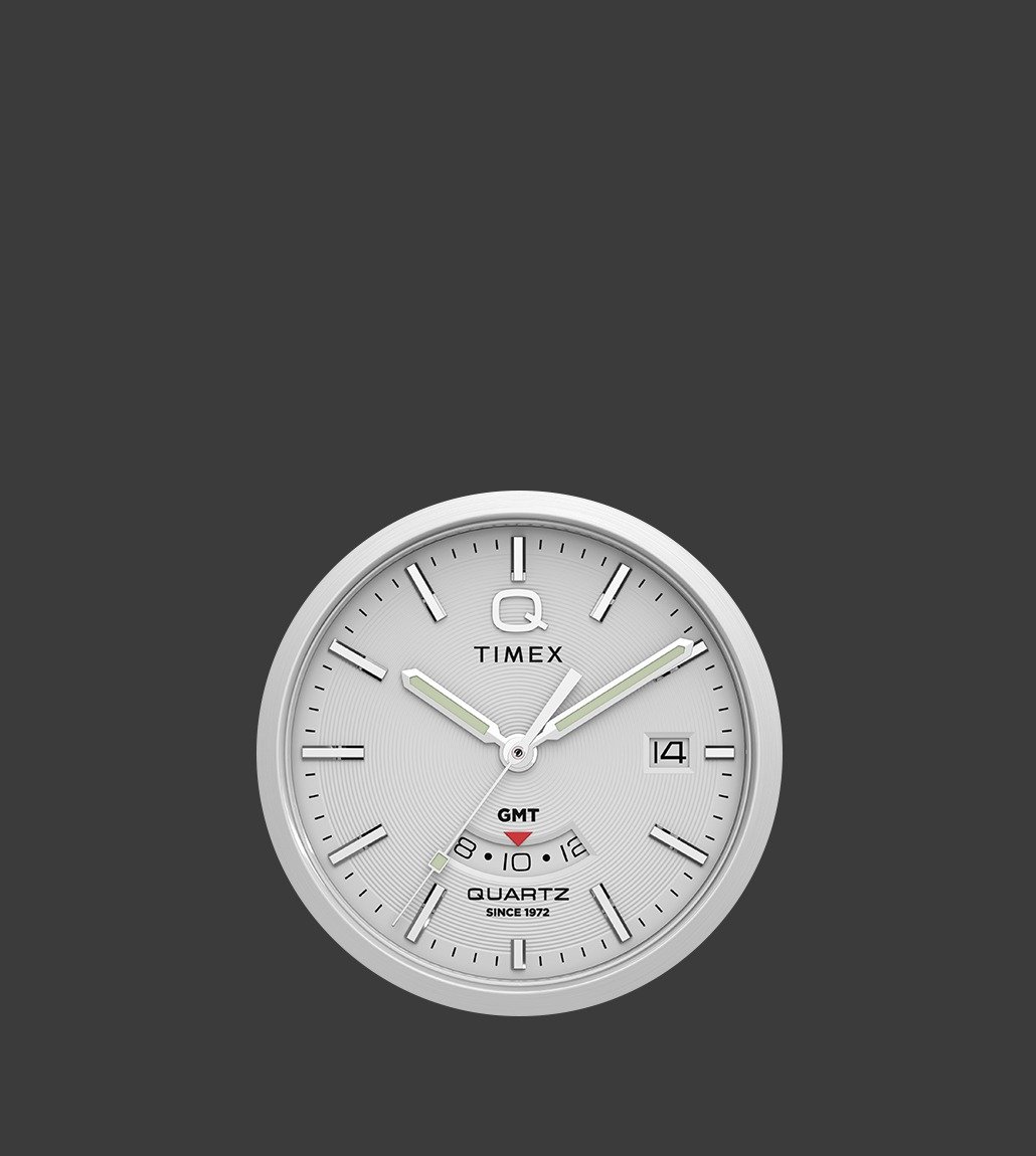 Close-up of the Q Timex® Continental GMT watch dial showcasing concentric dial texture, GMT sub-dial with red arrow hand, and polished hour markers against a silver background.