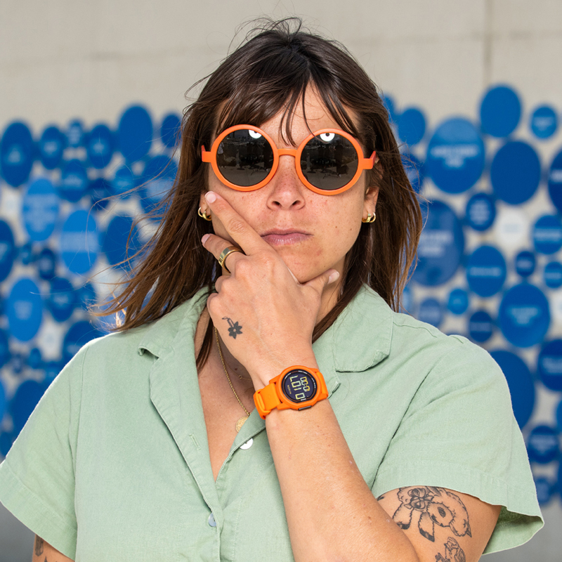 Nora Vasconcellos poses wearing orange sunglasses wearing an orange Nixon Disk Watch.