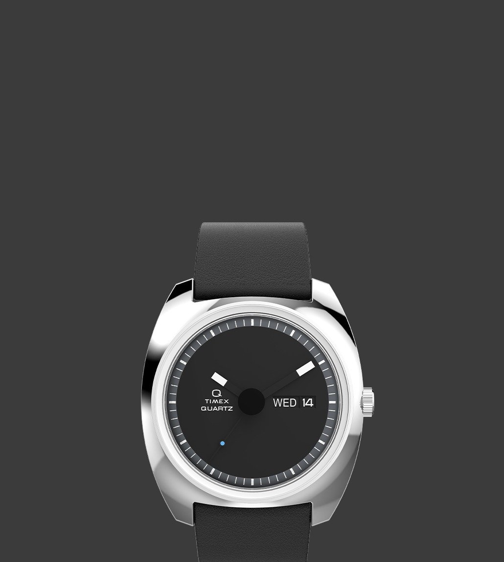 Front view of the Q Timex® 1975 Enigma watch showcasing a minimalist black dial, silver-tone case, bold white hour markers, and day-date display on the right side. The watch has a smooth black leather strap.